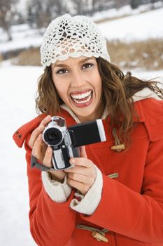 Caucasian young adult female in winter clothing holding video camera and smiling at viewer.