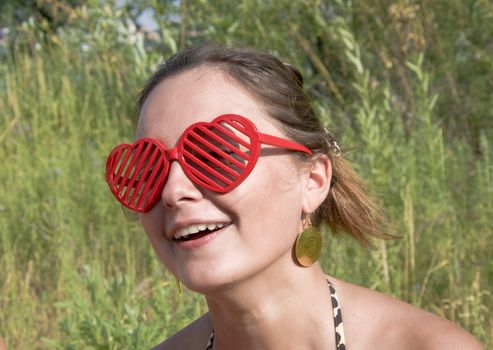 Beautiful young girl smiling in red sunglasses in the form of hearts. Summer portrait, close-up. Outdoor photoset.