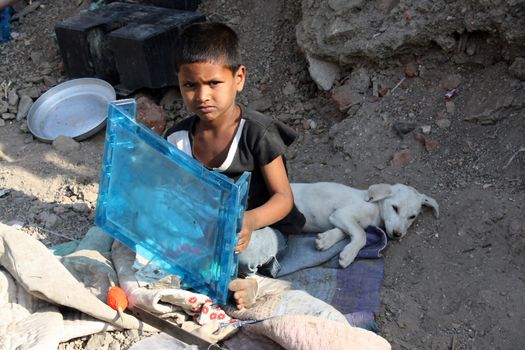 A poor boy sitting on the street-side with his white pet dog in India.