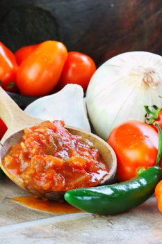 Rustic wooden spoon filled with spicy fresh salsa, surrounded by freshly picked and washed ingredients spilling from an old wooden bowl.