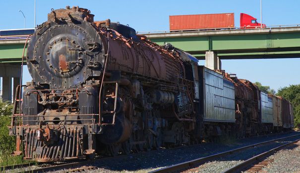 Old rusted train engine and cars against background of elevated highway with truck