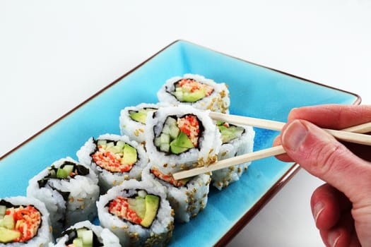 A hand holding sushi with a pair of chopsticks with more sushi in the background.