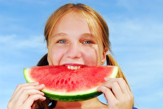 Young girl biting into a slice of watermelon on blue sky background