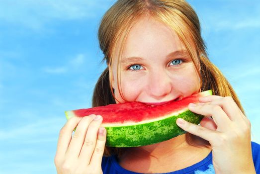 Young girl biting into a slice of watermelon on blue sky background