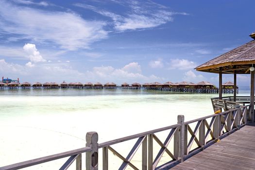 Image of a walkway and huts on stilts on a remote Malaysian tropical island with deep blue skies and crystal clear waters.