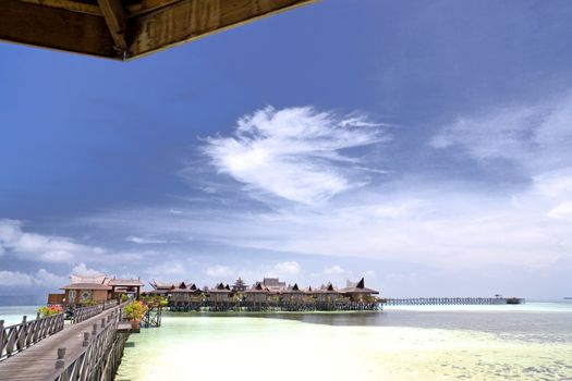 Image of a walkway and huts on stilts on a remote Malaysian tropical island with deep blue skies and crystal clear waters.