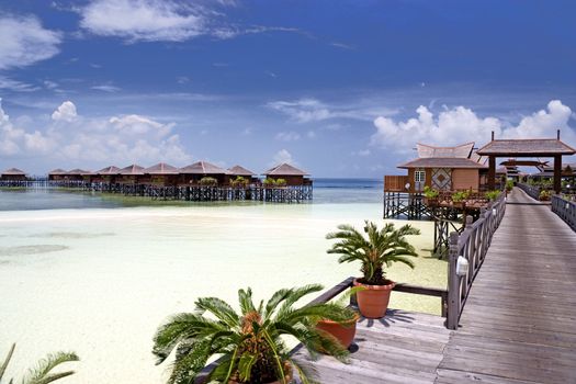Image of a walkway and chalets on stilts on a remote Malaysian tropical island with deep blue skies and crystal clear waters.