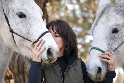 A pretty dark haired woman with two grey horses is kissing one of them on the muzzle.