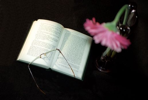 Open book and glasses on table with flower in vase 