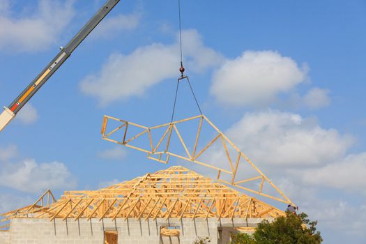 Roof Truss hanging from a crane being installed onto a new roof. Blue sky with clouds in background.
