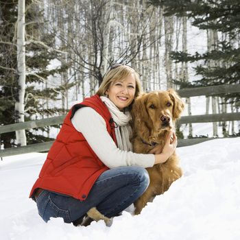 Attractive smiling mid adult Caucasian blond woman squatting in snow with arms around Golden Retriever.
