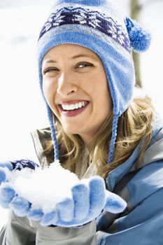 Portrait of attractive smiling mid adult Caucasian blond woman wearing blue ski cap and gloves holding snow towards viewer.