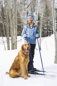 Mid adult smiling Caucasian female skier wearing blue ski clothing standing on ski slope with Golden Retriever.