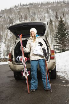 Attractive young woman in winter clothes standing holding ski equipment and smiling by automobile.