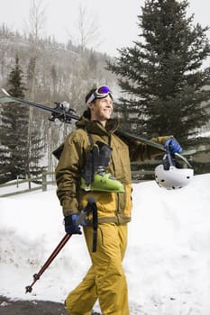 Attractive man in winter clothing walking in snow carrying ski equipment and smiling.