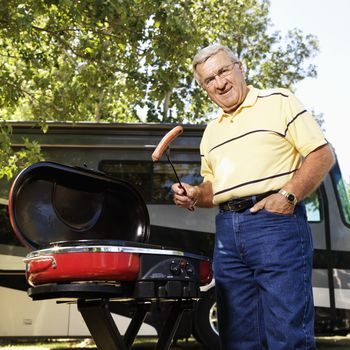 Senior adult man grilling hotdogs with RV in background.