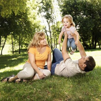 Caucasian mid adult parents holding toddler daughter in the air playing at grassy park.