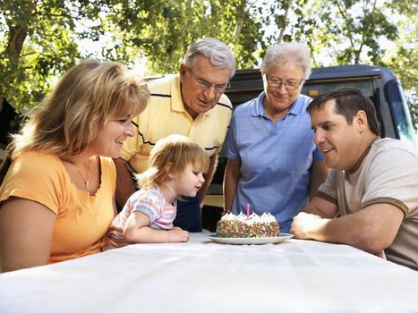 Three generation Caucasian family seated at picnic table celebrating female childs birthday with cake.