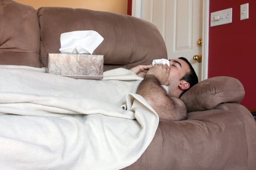 A young adult sick on the couch at home blows his nose with a tissue.