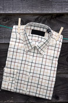 A gentelmens checked cotton shirt hanging on a washing line held by two wooden cloths pegs, set against a wooden building backdrop.