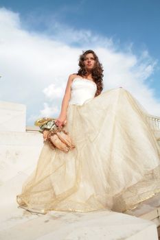 dark-haired princess in white-golden gown with doll in hand on step palace on background blue sky with white cloud