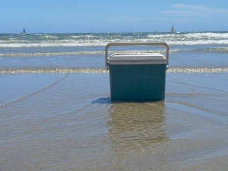 A cooler rests on the sand in the shallow water