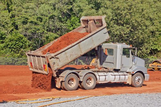 A tip truck is dumping red dirt or soil at a construction site