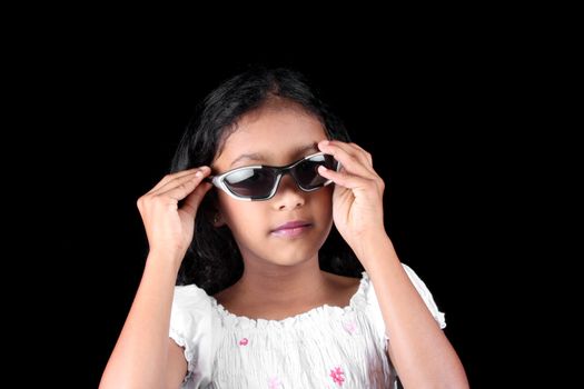 A portrait of a little Indian girl trying to be stylish by wearing trendy sunglasses, on black studio background.