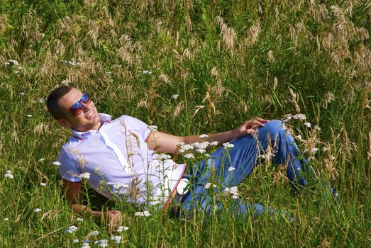 Young man in jeans with teeth smile is laying on summer field. Green grass surrounds him.