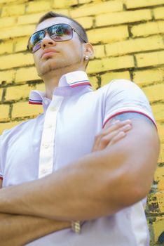 Man with big and muscled arms stands near the yellow brick wall. He wears sun glasses and earrings. Under the shirt you can see part of his tattoo.