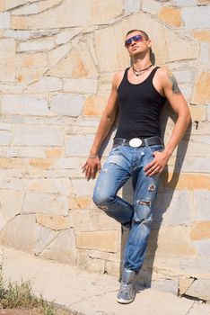 Person in sun glasses stands near the orange wall and looks at the sun. He has tattoo and earrings.