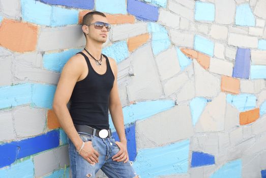 Young man in street wear and sun glasses stays with blank expression near the multi-colored wall. The city reflects in his glasses.