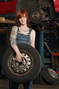 Photo of a young beautiful redhead mechanic wearing overalls and holding a wheel.  Attached property release is for arm tattoos.