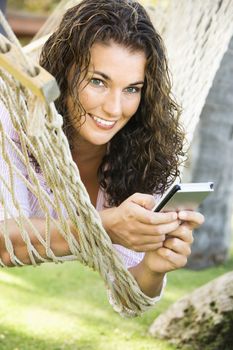 Smiling pretty young adult Caucasian brunette female lying in hammock using PDA.