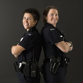 Portrait of two mid adult Caucasian policewomen standing back to back looking over their shoulders smiling at viewer.