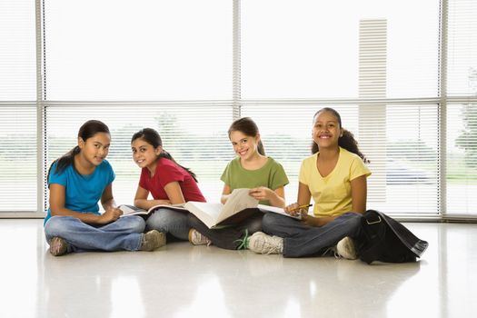 Preteen girls of mutiple ethnicities sitting together on floor with schoolwork smiling at viewer.