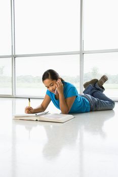 Asian preteen girl lying on floor doing homework while talking on cell phone.