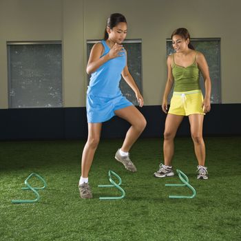 Girl jumping step hurdles in indoor gym while woman coach watches.