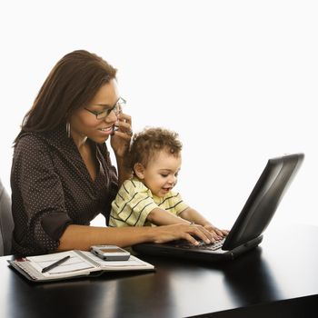 African American businesswoman at work on laptop and cell phone with toddler son on lap.