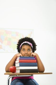 African American girl sitting in school desk with large stack of books looking bored.