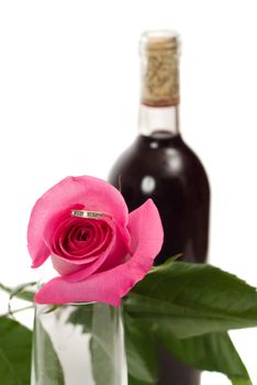 Closeup view of a ring hidden inside a rose, with an out of focus bottle of wine in the background, isolated against a white background