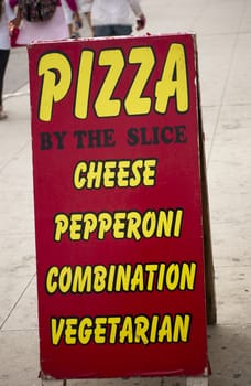 Pizza by the slice sign out on a busy sidewalk