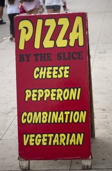 Pizza by the slice sign out on a busy sidewalk