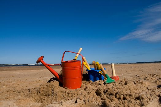 Red watering can, plastic blue bucket and other beach toys in the sandy seashore