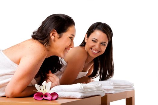Two beautiful women friends laying on wooden tables with towels waiting for their massage in the spa, smiling laughing talking and having fun, isolated.