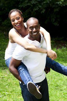 Beautiful happy smiling laughing African American couple piggyback playing in the park, woman hugging man, wearing white shirts and blue jeans.