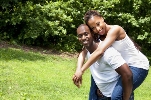 Beautiful happy smiling laughing young African American couple piggyback playing in the park, woman hugging man, wearing white shirts and blue jeans.