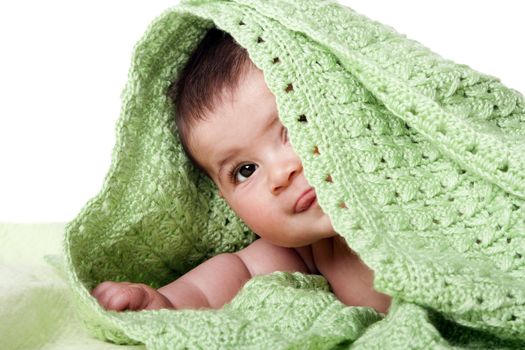 Beautiful cute happy Caucasian Hispanic baby face showing one eye and sticking out tongue while laying and hiding under covered between green knitted blankets, isolated.
