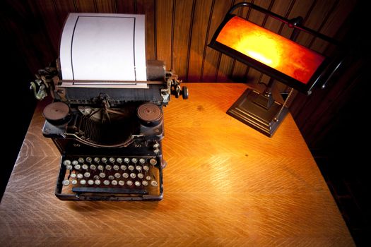 Wooden desk lit up by an old lamp with yellow light and a historic rusty typewriter with a plastified sheet of white paper.