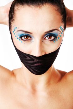 Beautiful female fashion face with blue makeup and gems rhinestones of a Caucasian Hispanic woman with hands behind head and bare shoulders and mouth covered with net-stockings, isolated.